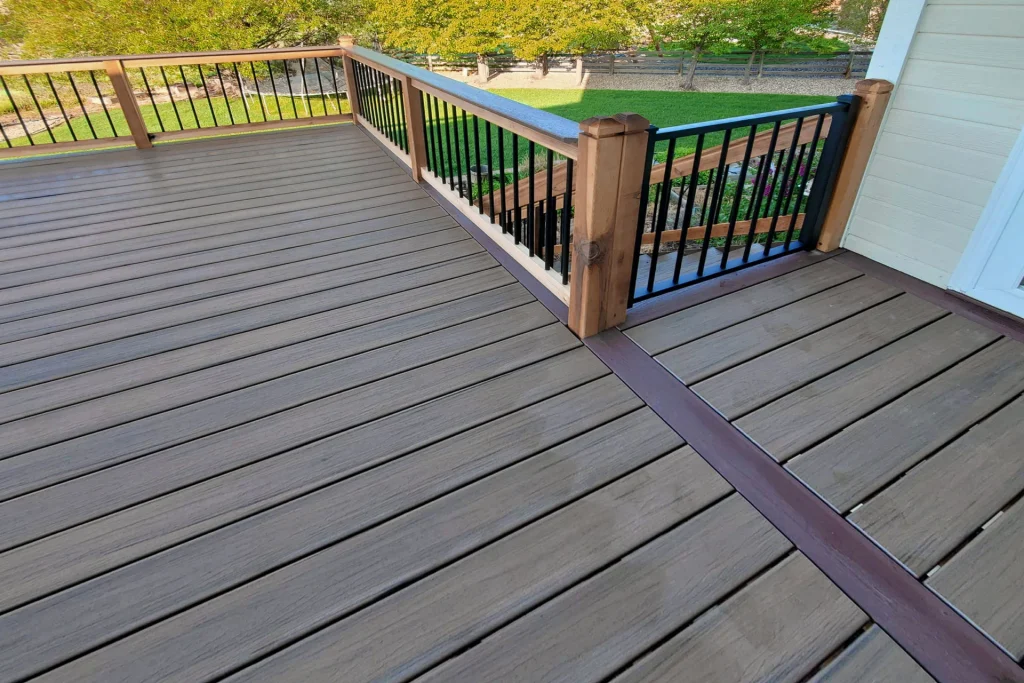 A spacious, elevated deck with brown composite flooring and a wooden railing featuring black metal balusters, overlooking a green lawn and trees in the background.