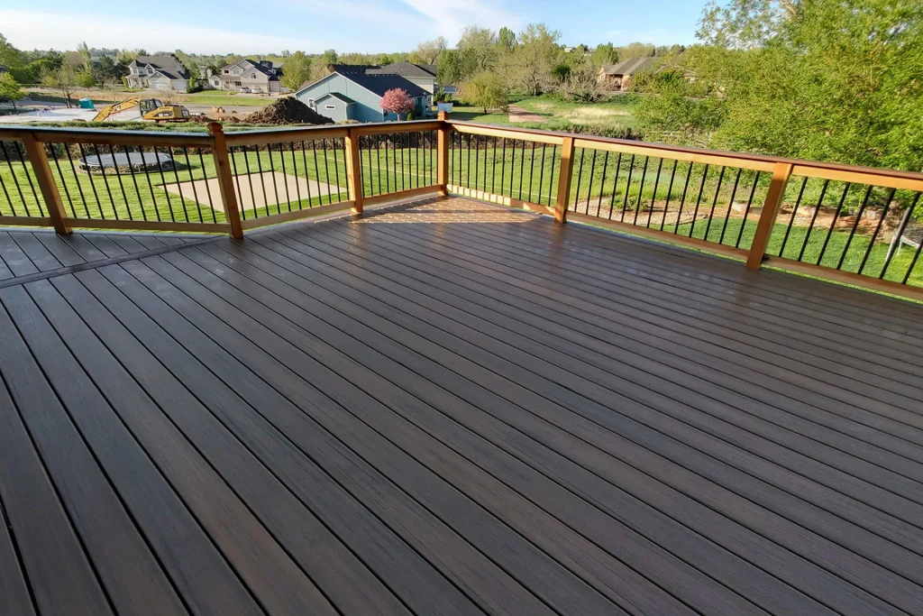 A spacious, empty wooden deck with a railing overlooks a green lawn, trees, and several houses in a suburban neighborhood under a clear sky.