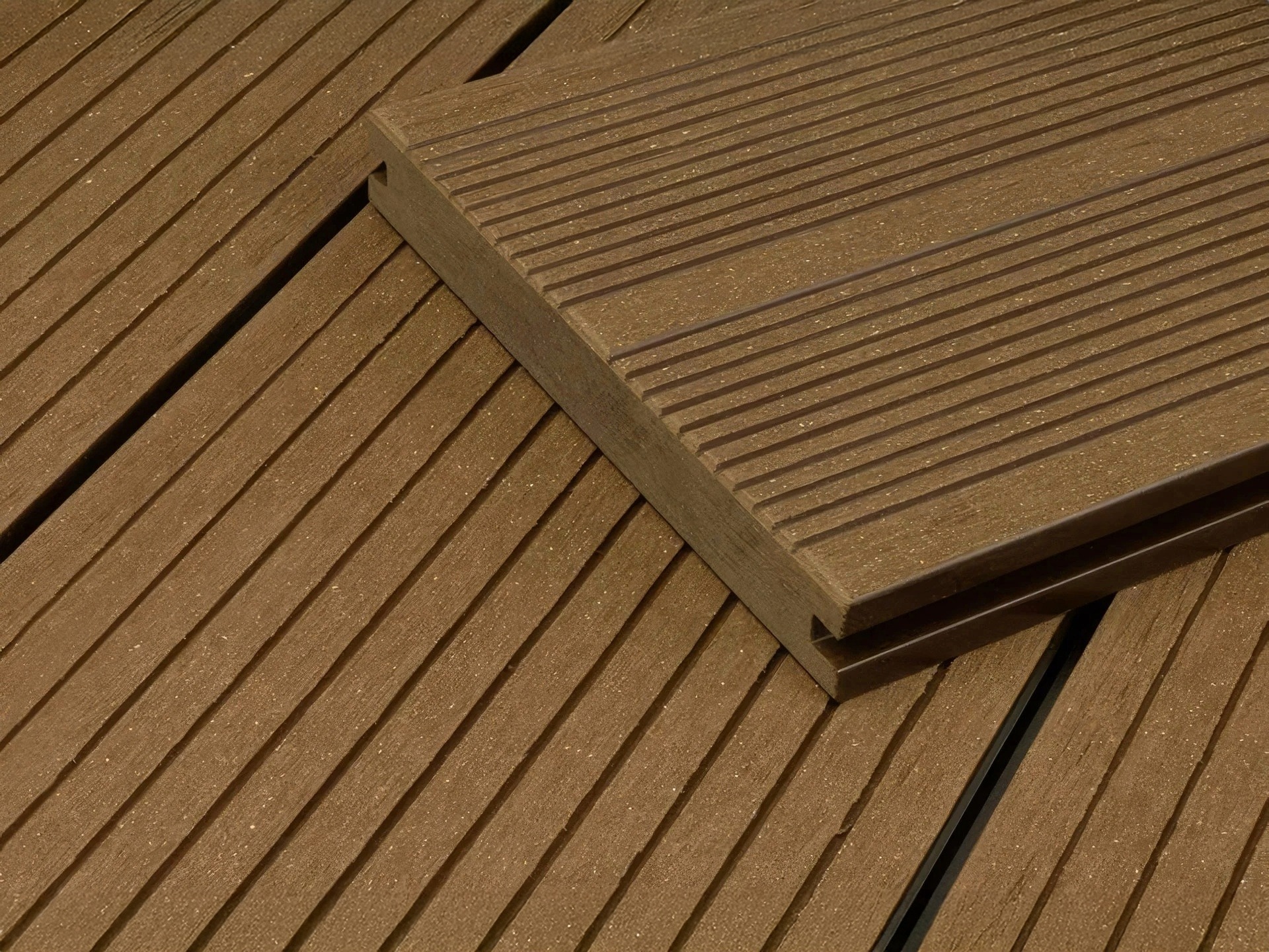 Close-up of brown composite decking boards with parallel grooves. One board is placed diagonally on top of others, showing its profile and textured surface. The decking has a uniform, modern appearance.