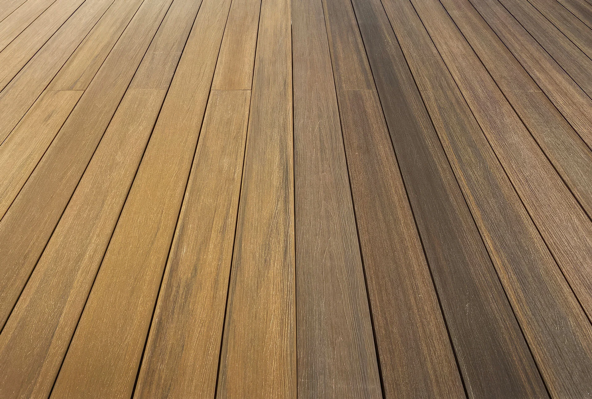 Close-up view of a wooden deck with long, narrow planks arranged parallel to each other. The boards display a gradient of colors from light brown on the left to dark brown on the right.
