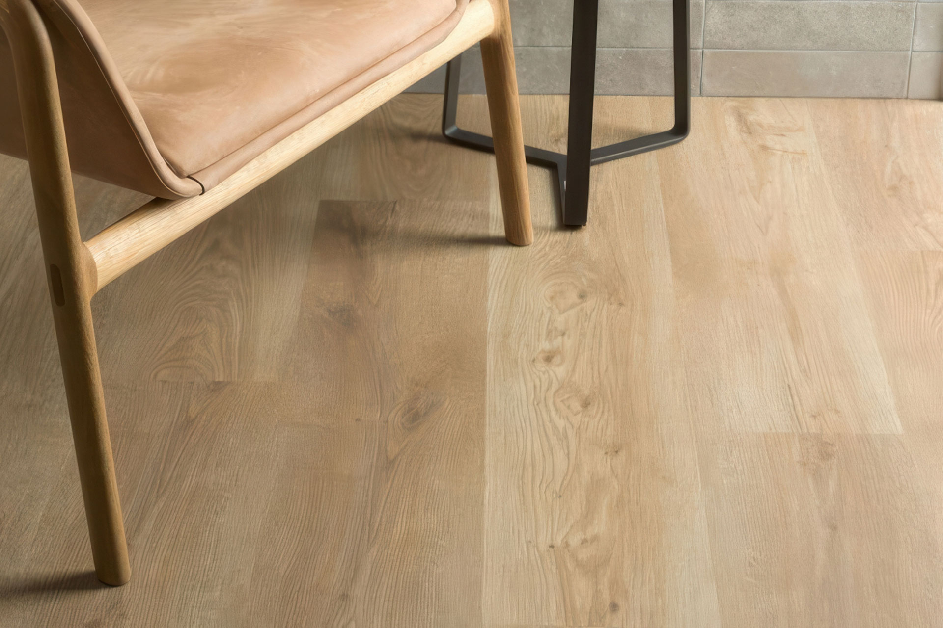 Close-up of a light wood floor with a tan leather chair with wooden legs and a metal-legged table in the background. The setting appears modern and minimalistic, with soft natural lighting.