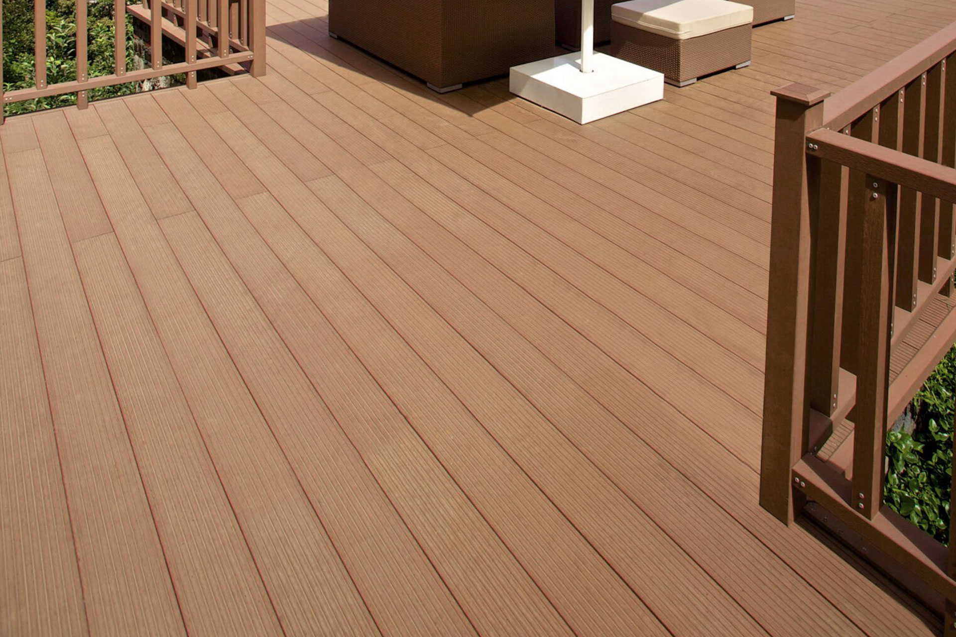 A close-up view of a clean, brown wooden deck with railings, featuring a few pieces of outdoor furniture and some greenery visible beyond the railing.