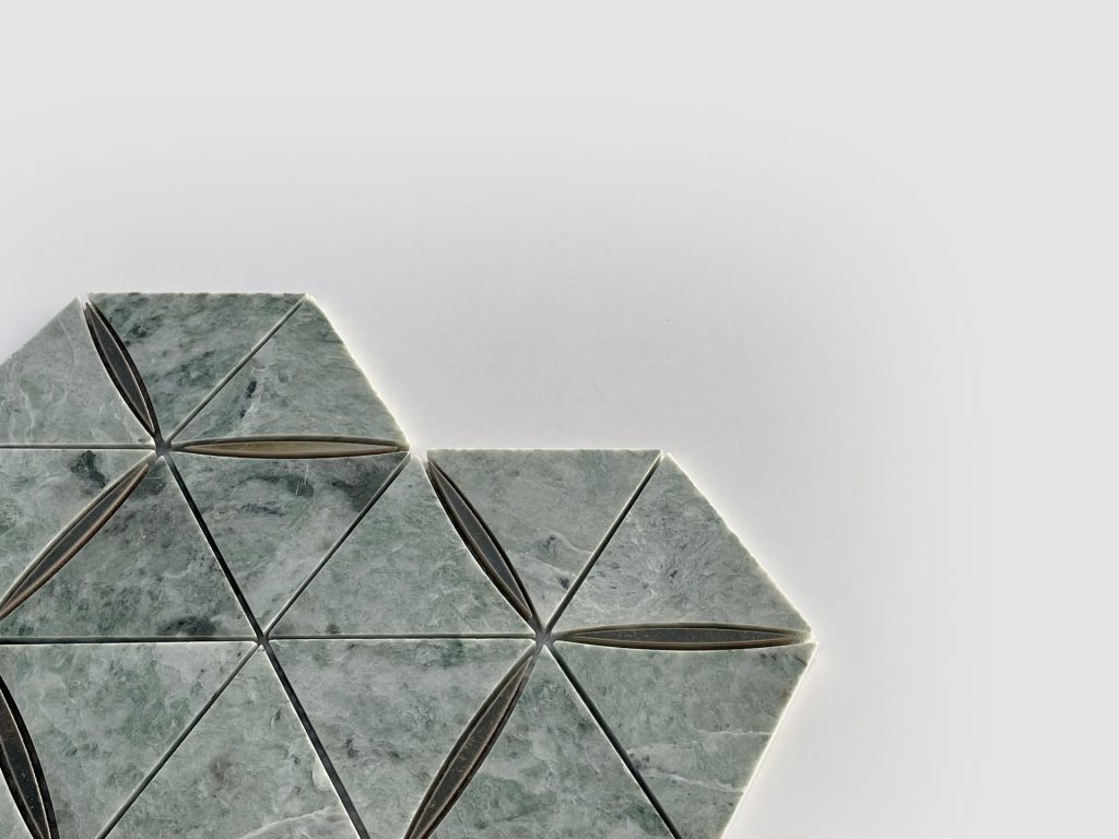 Geometric tile pattern with greenish-gray triangular tiles, arranged in a hexagonal formation on a white background. Some tiles have a thin, dark oval in the center. The design is partially shown, with tiles on the left side.