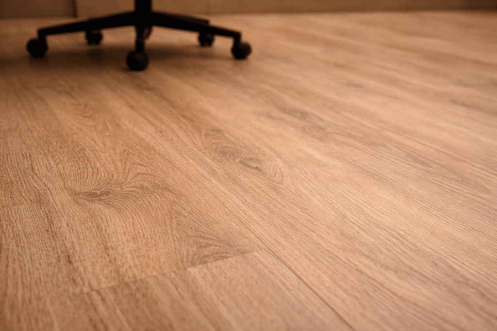 Close-up view of light brown wood-patterned flooring in an indoor room, with the base and wheels of a black office chair partially visible in the top left corner.