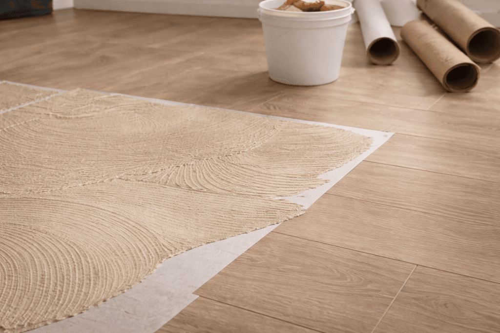 Close-up of flooring installation with adhesive spread on a sheet over wood-look tiles. In the background, there’s a white bucket and rolls of flooring material on the floor.