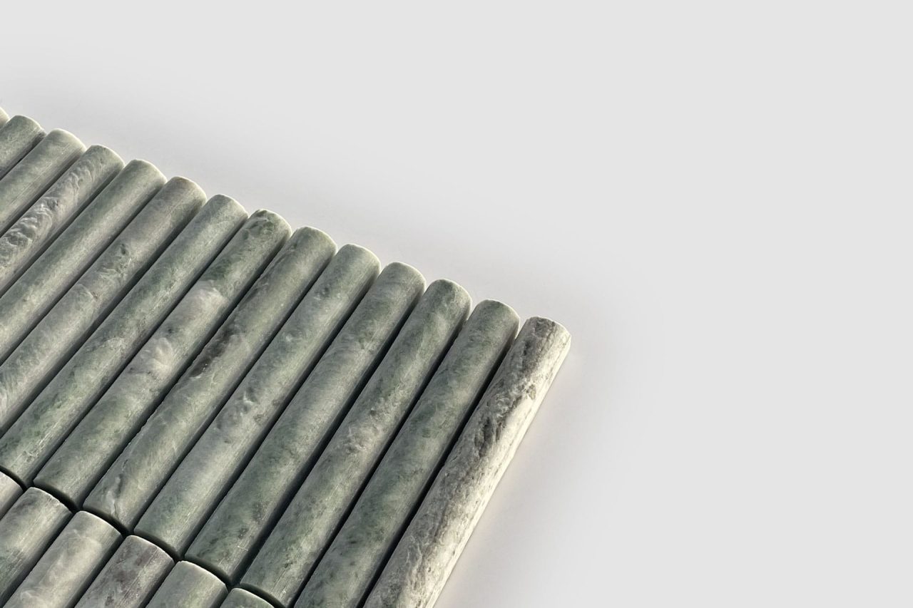 A close-up of evenly spaced, cylindrical green marble rods arranged in parallel rows on a light gray background. The rods have a smooth, polished surface with natural marbling patterns.