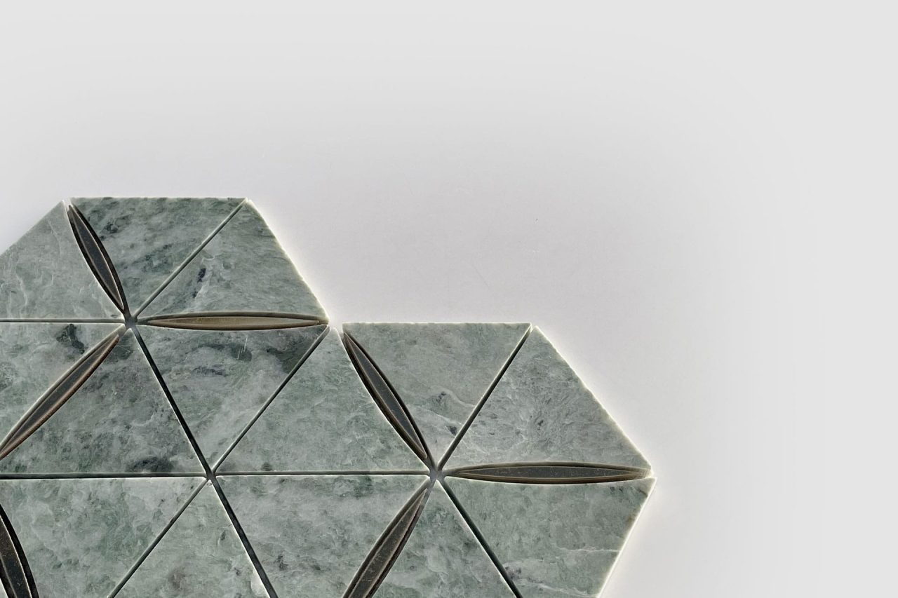 Geometric tile pattern with greenish-gray triangular tiles, arranged in a hexagonal formation on a white background. Some tiles have a thin, dark oval in the center. The design is partially shown, with tiles on the left side.